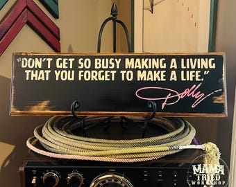 May include: A rectangular wooden sign with a black finish and distressed brown edges. The text reads, "DON'T GET SO BUSY MAKING A LIVING THAT YOU FORGET TO MAKE A LIFE." with the name "Dolly" in pink script lettering on the lower right side.