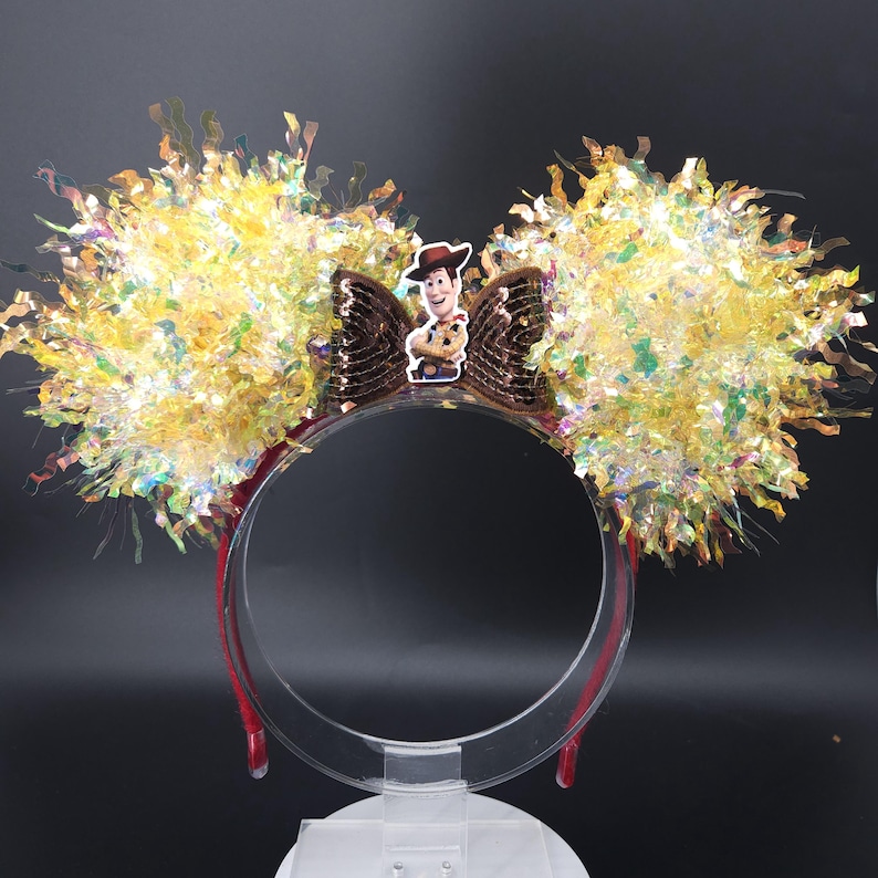 Puede incluir: Una diadema festiva con grandes orejas amarillas y brillantes. Un lazo de lentejuelas marr&oacute;n con una imagen del personaje de Woody est&aacute; centrado en la diadema. La diadema es roja y de pl&aacute;stico transparente.
