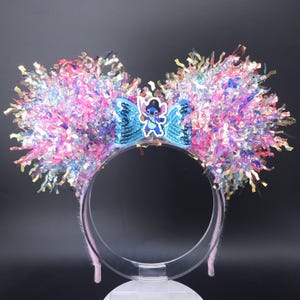May include: A pair of mouse ears with a clear headband. The ears are decorated with pink, blue, and iridescent tinsel. A blue sequined bow features a Stitch character. The headband has pink velvet padding.