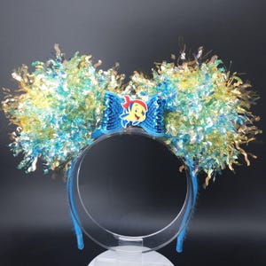 May include: A blue headband with large, fluffy ears. The ears are a mix of blue, yellow, and gold tinsel. A blue bow with a cartoon character is centered on the headband. The headband is clear plastic.