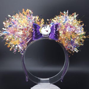 May include: Purple headband with a large purple bow and iridescent gold and purple fringe. The bow has a character from Beauty and the Beast, Mrs. Potts, on it.