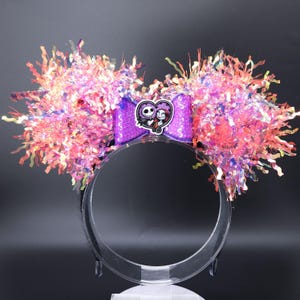 Puede incluir: Orejas de Minnie Mouse iridiscentes de color morado y rosa con un lazo morado y un amuleto de Jack y Sally.