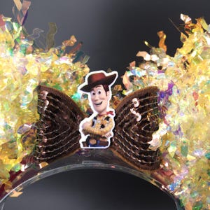 Puede incluir: Una diadema transparente con un lazo de lentejuelas marr&oacute;n con un recorte del personaje de Woody. La diadema est&aacute; decorada con confeti dorado e iridiscente, creando un aspecto festivo y juguet&oacute;n. Perfecto para fiestas tem&aacute;ticas o cosplay.