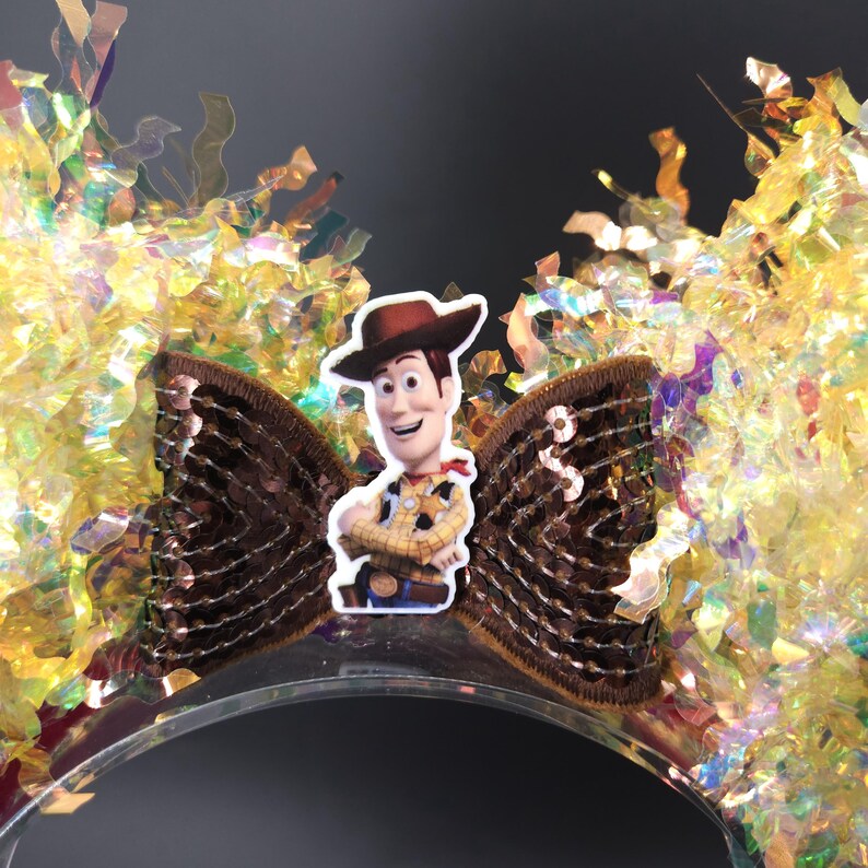 Puede incluir: Una diadema transparente con un lazo de lentejuelas marr&oacute;n con un recorte del personaje de Woody. La diadema est&aacute; decorada con confeti dorado e iridiscente, creando un aspecto festivo y juguet&oacute;n. Perfecto para fiestas tem&aacute;ticas o cosplay.