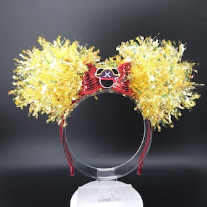 May include: A decorative headband featuring Mickey Mouse ears. The ears are made of gold tinsel and a red sequined bow with a pirate hat emblem. The headband is red and clear.