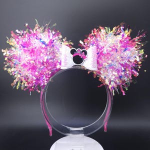 May include: A decorative headband featuring large, fluffy ears made of pink and iridescent tinsel. A white bow with a Minnie Mouse silhouette is centered on the headband. The headband is clear with pink accents.