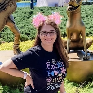 Puede incluir: Una mujer con una camiseta negra con el texto "Epcot Food & Wine Festival" y orejas rosas brillantes. Sonr&iacute;e frente a una estatua dorada de un perro y una estatua de un m&uacute;sico. El fondo incluye vegetaci&oacute;n y un lago.