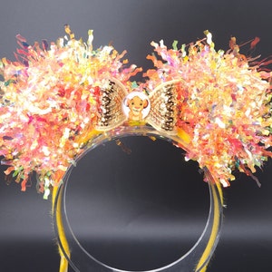 May include: A yellow headband with a large bow and a sparkly, iridescent, orange and pink pom-pom on each side. The bow has a small, round, gold button with a picture of Simba from The Lion King.