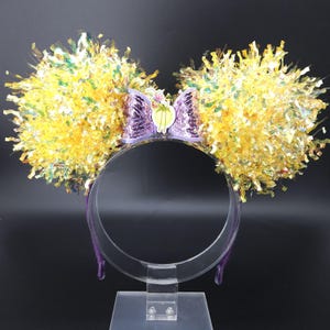 May include: A purple headband with yellow and green iridescent pom-poms on each side. A purple sequin bow with a green and yellow flower charm is in the center.