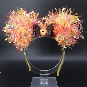 May include: A festive headband with large, fluffy ears made of orange, yellow, and iridescent tinsel. A brown sequined bow sits between the ears, with a character button in the center. The headband is yellow and clear.