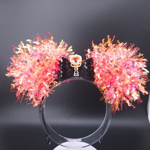 May include: Black headband with a large black bow and a cartoon character charm. The bow is covered in black sequins and the headband is decorated with bright orange and pink iridescent tinsel.