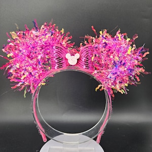Puede incluir: Diadema rosa con orejas grandes y esponjosas hechas de oropel y lentejuelas rosas. Un lazo rosa con lentejuelas y una pequeña cabeza de Mickey Mouse rosa claro está centrado en la diadema. La diadema es de plástico transparente.