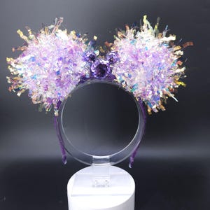 May include: A purple headband with large, fluffy ears. The ears are adorned with iridescent, glittery fringe and a central Mickey Mouse-shaped jewel. The headband is displayed on a clear stand against a dark background.