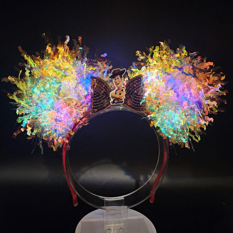 Puede incluir: Una diadema roja con orejas grandes e iluminadas. Las orejas est&aacute;n adornadas con flecos iridiscentes y coloridos y un lazo marr&oacute;n con una imagen del personaje Woody. La diadema est&aacute; sostenida por un expositor transparente.