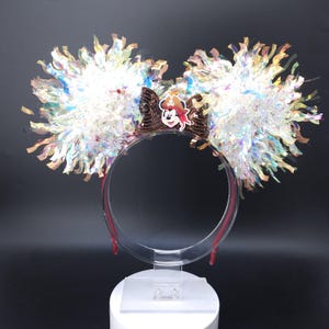 May include: Mouse ear headband featuring large, fluffy, iridescent pom-poms. The clear headband has a red accent. A brown bow with a Mickey Mouse pirate graphic is centered. The ears are displayed on a clear stand.