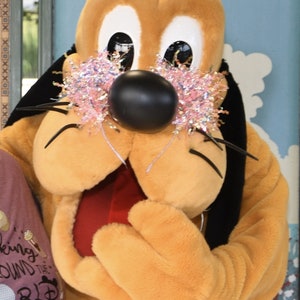 Puede incluir: Un disfraz del personaje de Pluto con una gran nariz negra, ojos blancos y una decoraci&oacute;n rosa brillante alrededor de los ojos. El disfraz es de color canela y amarillo con orejas negras y una lengua roja. El fondo es una pared azul claro con dise&ntilde;os florales blancos.