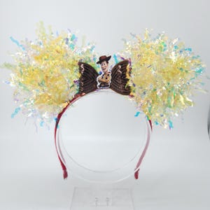Puede incluir: Una diadema roja con un lazo marr&oacute;n con una imagen del personaje Woody. El lazo est&aacute; flanqueado por grandes acentos de oropel amarillo e iridiscente, creando un aspecto festivo. Ideal para eventos tem&aacute;ticos o fiestas de disfraces.