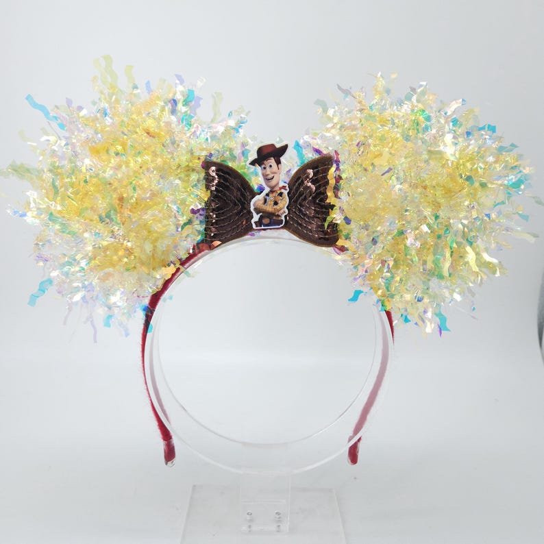 Puede incluir: Una diadema roja con un lazo marr&oacute;n con una imagen del personaje Woody. El lazo est&aacute; flanqueado por grandes acentos de oropel amarillo e iridiscente, creando un aspecto festivo. Ideal para eventos tem&aacute;ticos o fiestas de disfraces.