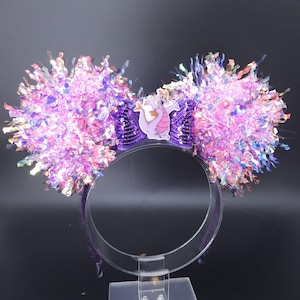 May include: Purple and iridescent ear headband with large, fluffy, pom-pom-like ears. A purple sequined bow features a cartoon dragon. The headband is a fun accessory for theme park enthusiasts.