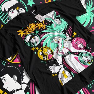 Puede incluir: Camiseta negra con un estampado gráfico colorido que presenta personajes de anime y el texto "天 地 無 用!" en japonés. El diseño incluye estrellas, remolinos y círculos.