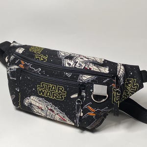 Puede incluir: Riñonera negra con temática de Star Wars. La riñonera presenta un patrón repetido de naves espaciales y el logotipo de Star Wars en amarillo. Tiene múltiples compartimentos con cremallera y una correa negra ajustable.