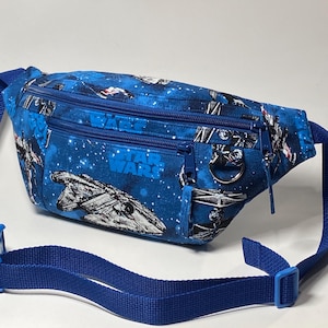 May include: A blue fanny pack featuring a Star Wars space print with various starships and the Star Wars logo. The bag includes two zippered compartments, a metal D-ring attachment, and an adjustable blue nylon strap with a plastic buckle closure.