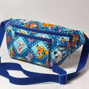 May include: A blue fanny pack featuring a repeating pattern of various cartoon dogs in diamond-shaped frames. The bag has two zippered compartments, a blue adjustable strap with a plastic buckle, and a silver metal D-ring attached to the front.