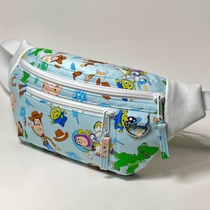 May include: A light blue fanny pack featuring a pattern of cartoon characters. The bag has a white strap and white accents. It has two zippered compartments with silver zippers and a small charm.