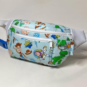 May include: A light blue fanny pack featuring a repeating pattern of Toy Story characters including Woody, Buzz Lightyear, Rex, and green aliens. The bag has white zippers, white side panels, a white adjustable strap, and a silver metal D-ring.