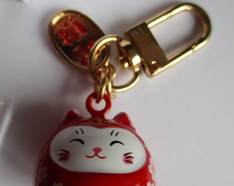 May include: A red and white Daruma doll keyring with a gold-coloured clasp and accents. The doll features a smiling face and Japanese characters. Floral designs adorn the sides of the doll.
