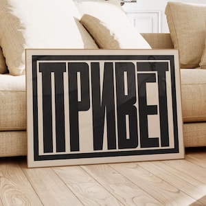 Puede incluir: Un póster en blanco y negro con la palabra "ПРИВЕТ" en letras negritas y en bloque. El póster está enmarcado y colgado en una pared sobre un sofá beige con almohadas.