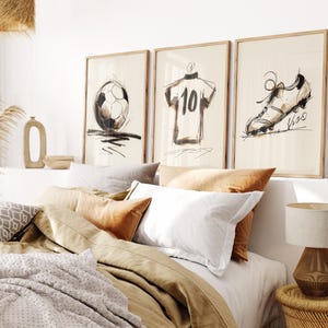 Puede incluir: Tres impresiones artísticas enmarcadas con bocetos de elementos relacionados con el fútbol. Las impresiones representan un balón de fútbol, una camiseta con el número 10 y una bota de fútbol. La obra de arte es en blanco y negro con un estilo minimalista, sobre un fondo neutro.