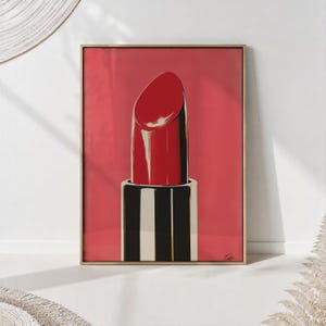 Op de afbeelding: Een ingelijste kunstdruk met een gestileerde illustratie van een felrode lippenstift tegen een effen rode achtergrond. De lippenstift heeft een glanzende afwerking met witte accenten en de basis is uitgevoerd in afwisselende zwarte en witte strepen.