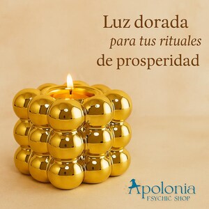 以下が含まれることがあります： 球状のデザインが特徴の、金色のキャンドルホルダーと点灯したキャンドル。テキストは「Luz dorada para tus rituales de prosperidad」と表示され、ショップ名は「Apolonia Psychic Shop」です。