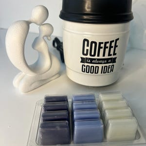 Puede incluir: Una taza de café de cerámica blanca con tapa negra y el texto "COFFEE is always a GOOD IDEA". También hay una escultura blanca de dos figuras abrazándose y una bandeja de ceras aromáticas en tonos azules y blancos.
