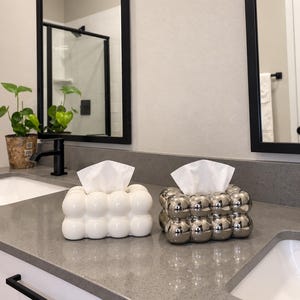 May include: Two rectangular tissue box covers with a bubble-like, spherical texture. One cover is finished in a smooth, glossy white, while the other features a reflective, metallic silver finish. Both items hold white tissues extending from the top.