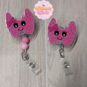 May include: Two badge reels featuring a pink thyroid-shaped character with a smiling face, eyelashes, and a small pink bow. One reel includes two round beads in light and dark pink. Above them is a pink scalloped logo reading The Shimmer Studio.