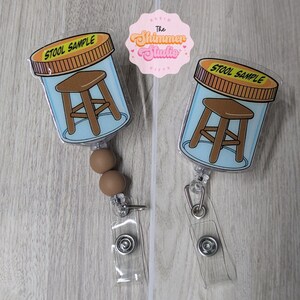 May include: Two badge reels featuring a clear plastic container graphic with a brown stool illustration inside and a yellow lid labeled STOOL SAMPLE. One reel includes two brown silicone beads above the clear plastic strap and metal clip.