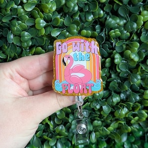 May include: A pink flamingo float with the text "Go with the float" on a gold glitter background. The float is surrounded by a rainbow and waves. The badge is attached to a clear retractable badge holder.