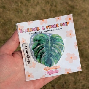 May include: A magnetic e-reader and phone grip featuring a green monstera leaf design. The product is packaged on a white card with orange flower illustrations, the text E-READER & PHONE GRIP, and the brand name Shimmer Studio in orange.