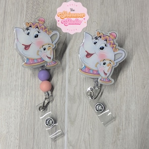 May include: Two badge reels featuring a cartoon teapot and teacup character design. The characters are white with pink, blue, and yellow accents. One reel includes two round beads in lavender and peach. Both have a clear plastic strap and metal snap.