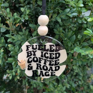 May include: A white round car air freshener with black text that reads "Fueled by Iced Coffee & Road Rage". A small cartoon image of a glass of iced coffee is next to the text. The air freshener is hanging from a black string with two wooden beads.