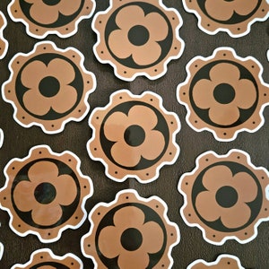 May include: A set of ten brown and black flower stickers. Each sticker features a stylized flower with five petals and a black center. The stickers are arranged in a circular pattern on a brown surface.