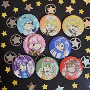 May include: A collection of round keychains featuring anime-style character illustrations. Each keychain has a different character with unique hair colors and outfits. The keychains are attached to silver star-shaped keyrings, set against a black background with star decorations.