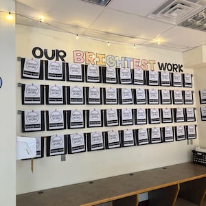 Könnte beinhalten: Eine Präsentation an der Wand eines Klassenzimmers mit den Worten "OUR BRIGHTEST WORK" in großen, farbigen Buchstaben. Darunter hängen zahlreiche schwarz-weiße Schilder mit dem Text "BRIGHT WORK" und "coming soon". Die Präsentation befindet sich über einem langen Schreibtisch.