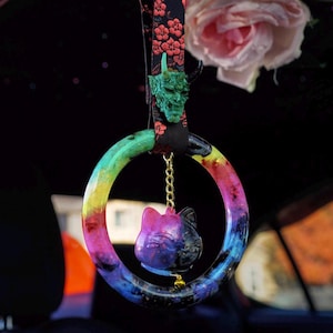 Puede incluir: Un anillo de resina de colores arcoíris con un dije de gato negro y morado colgando. El anillo está unido a una correa de tela negra con un dije de cara de demonio verde.