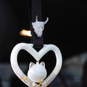 Peut inclure: Ornement en forme de cœur blanc avec une petite figurine de chat blanc à l'intérieur. L'ornement est décoré de paillettes dorées et est suspendu à une sangle en cuir noir avec un charme de tête de démon blanc.