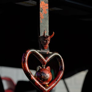 Puede incluir: Un amuleto de coche en forma de corazón rojo con una figurita de gato negro en su interior. El amuleto cuelga de una cinta negra con un patrón floral rojo. Una cabeza de demonio roja está unida a la parte superior de la cinta.