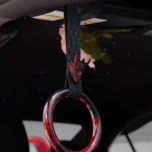 Puede incluir: Un accesorio de interior de coche rojo y negro con un amuleto de cabeza de demonio. El anillo está hecho de plástico transparente con purpurina roja incrustada.