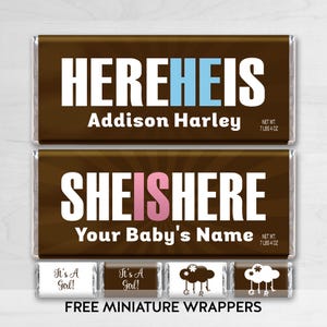 May include: Two brown chocolate bar wrappers with white text. The top wrapper says "HEREHEIS Addison Harley" and the bottom wrapper says "SHEISHERE Your Baby's Name".  The wrappers are for a baby shower.